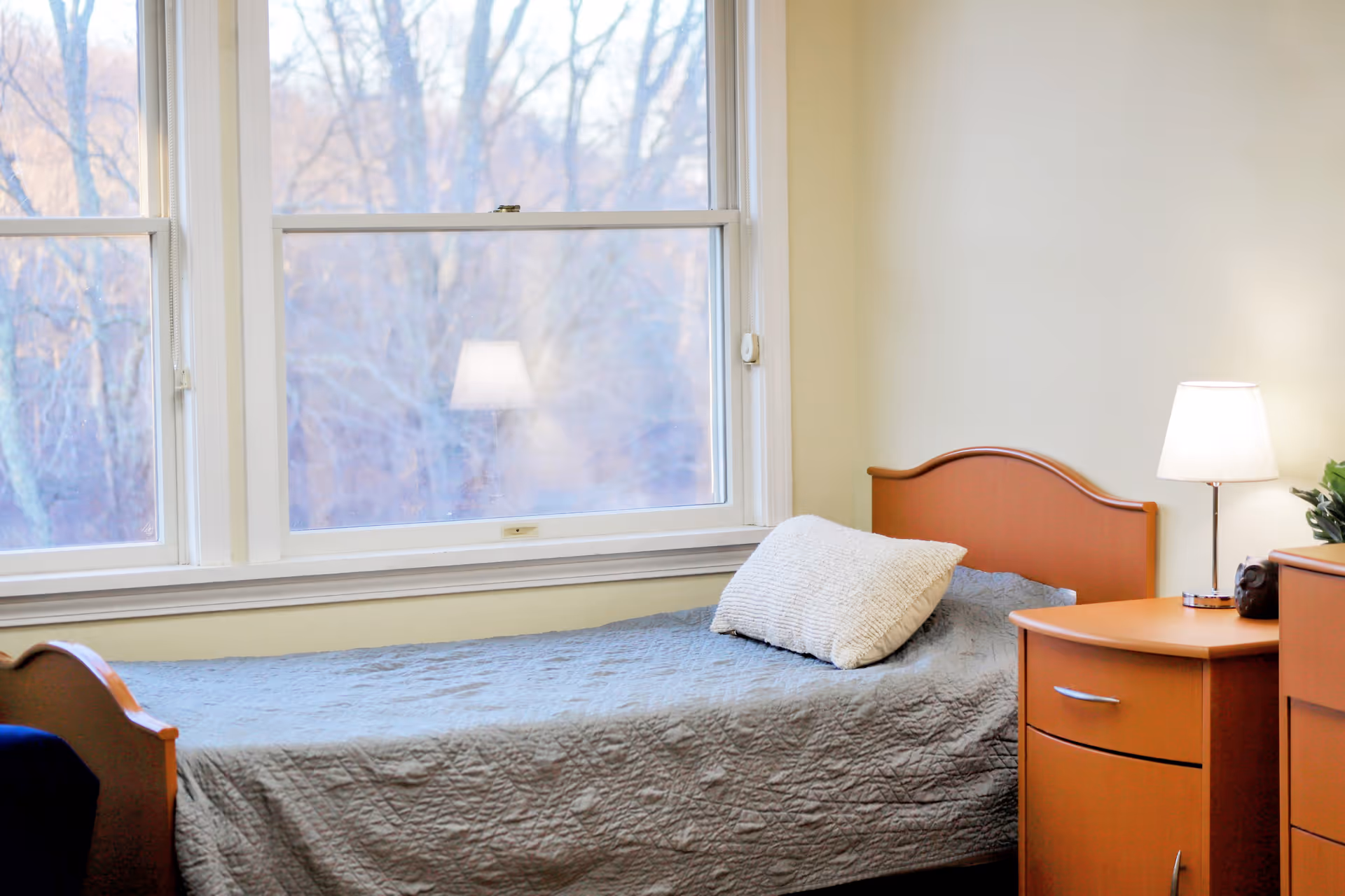 A simple bedroom with a single bed covered in a gray quilt, a pillow, a wooden headboard and nightstand with a lamp beneath large windows.