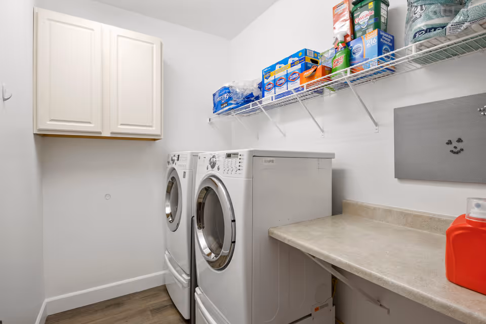 Laundry room with a front-loading washing machine and dryer side by side. Above them is a wire shelf stocked with various cleaning supplies and paper products. There is a beige countertop to the right and a white cabinet mounted on the wall to the left. The room has white walls and wood flooring.