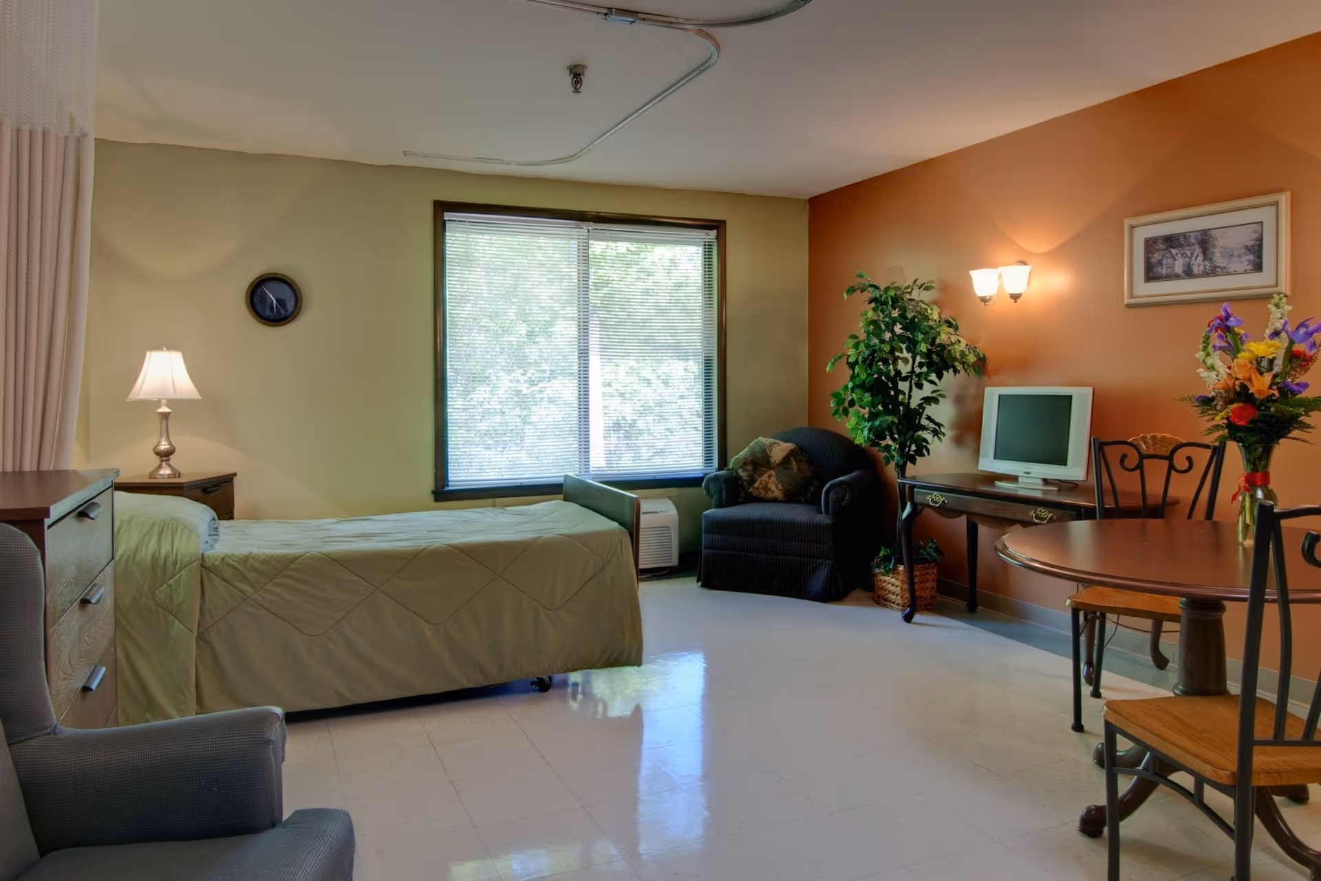 A nursing facility room with a single bed covered in a green bedspread, a bedside table with a lamp, a window with blinds, a comfortable armchair, a small table with a vintage TV, a potted plant, a round dining table with chairs, and a vase of flowers on the table. The walls are painted in light green and orange tones.