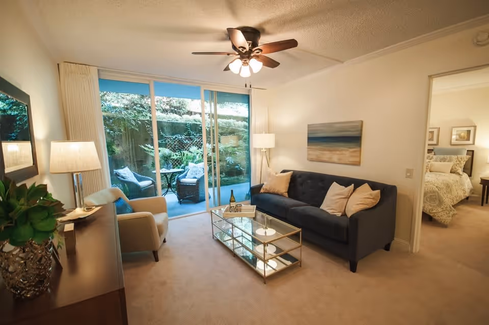A cozy living room with a dark blue sofa adorned with light-colored pillows, a glass coffee table with a bottle and tray on it, a beige armchair, a floor lamp, and a ceiling fan with lights. Large sliding glass doors open to a small outdoor patio with two wicker chairs and a table, surrounded by greenery. A doorway leads to a bedroom with a bed and nightstand visible.