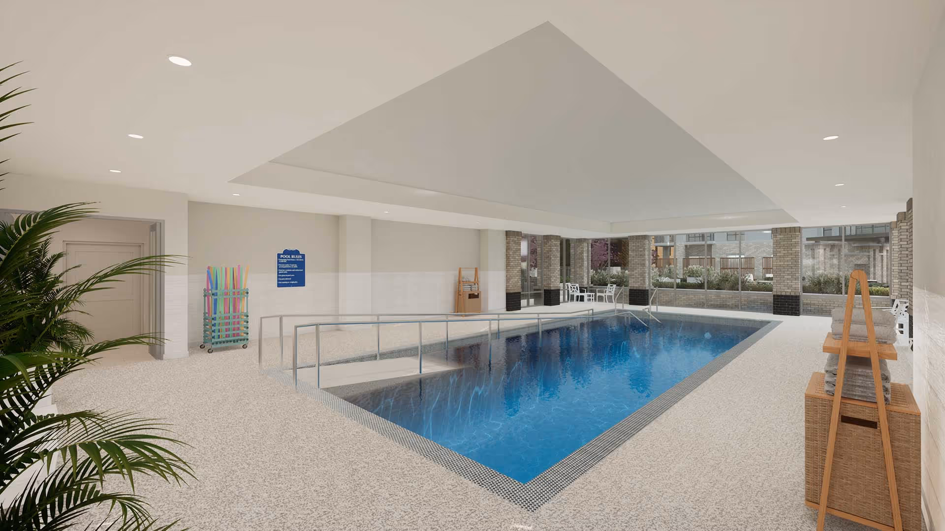 Indoor swimming pool area with a ramp for accessibility, surrounded by beige tiled flooring and walls. There are pool noodles stored on a rack near a door, a sign with pool rules on the wall, and a seating area with white chairs and tables near large windows showing an outdoor garden. Towels are neatly stacked on a wooden rack to the right.