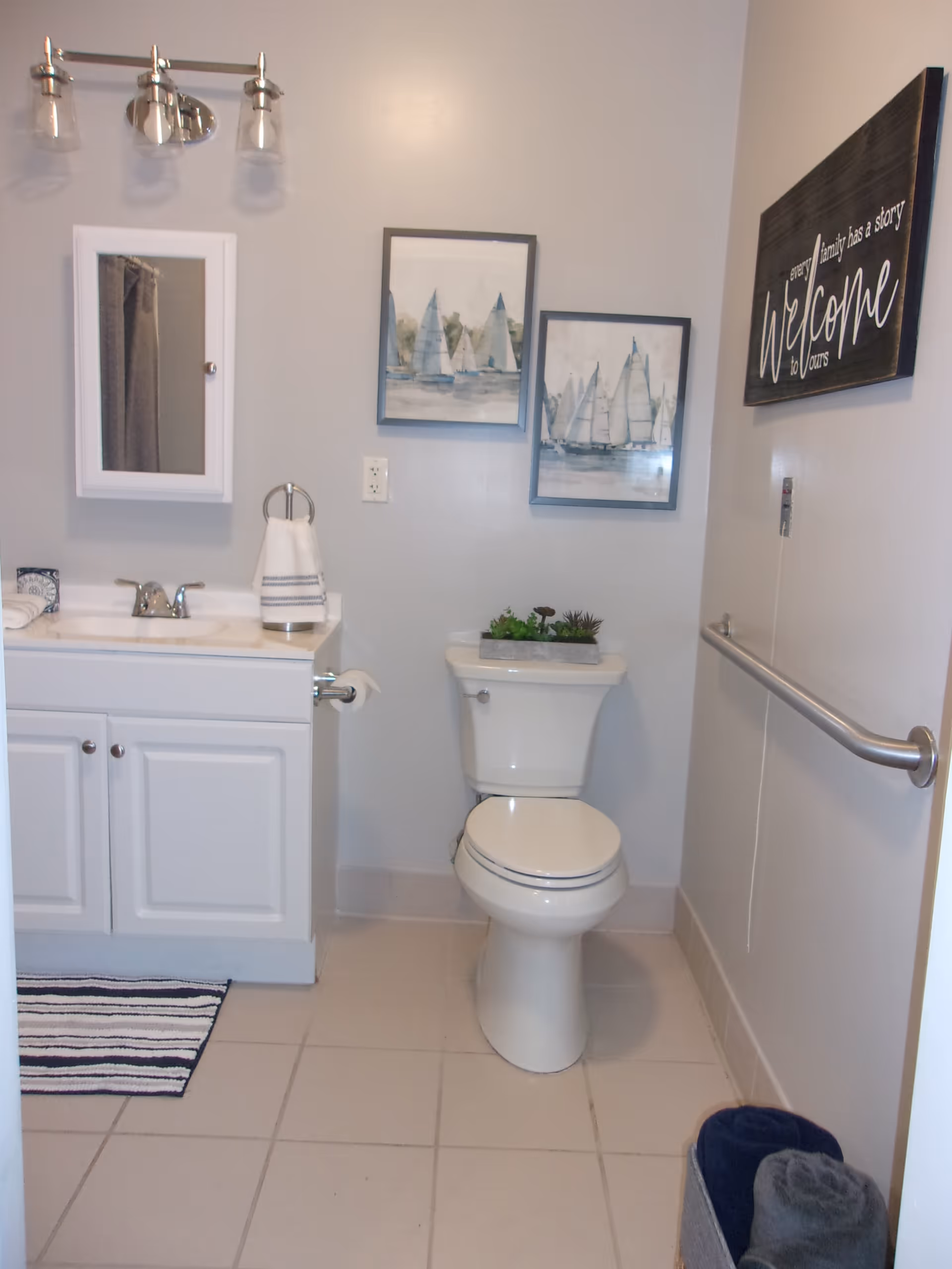 A clean bathroom with a white toilet and a white vanity with a sink. Above the vanity is a small mirror cabinet and a light fixture with three bulbs. Two framed pictures of sailboats hang on the wall above the toilet. A decorative plant is placed on the toilet tank. A towel ring with a hand towel is mounted next to the vanity. On the right wall, there is a grab bar and a sign that reads 'Every family has a story Welcome to yours.' A striped rug is on the tiled floor, and a basket with rolled towels is in the corner.