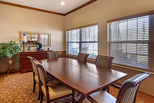 A dining room with a long wooden table surrounded by six chairs with striped upholstery. The room has beige walls with a wooden chair rail and crown molding. There are two large windows with blinds letting in natural light. Against one wall is a wooden sideboard with a mirror above it, a lamp, and a coffee maker. A potted plant is in the corner of the room.