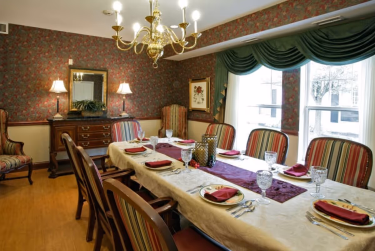 A formal dining room with a long table set for eight people. The table is covered with a white tablecloth and a purple runner, with plates, silverware, crystal glasses, and folded red napkins at each place setting. The chairs have striped upholstery. The room has floral wallpaper, a large window with green drapes, a chandelier, two table lamps on a wooden sideboard, and framed artwork on the walls.