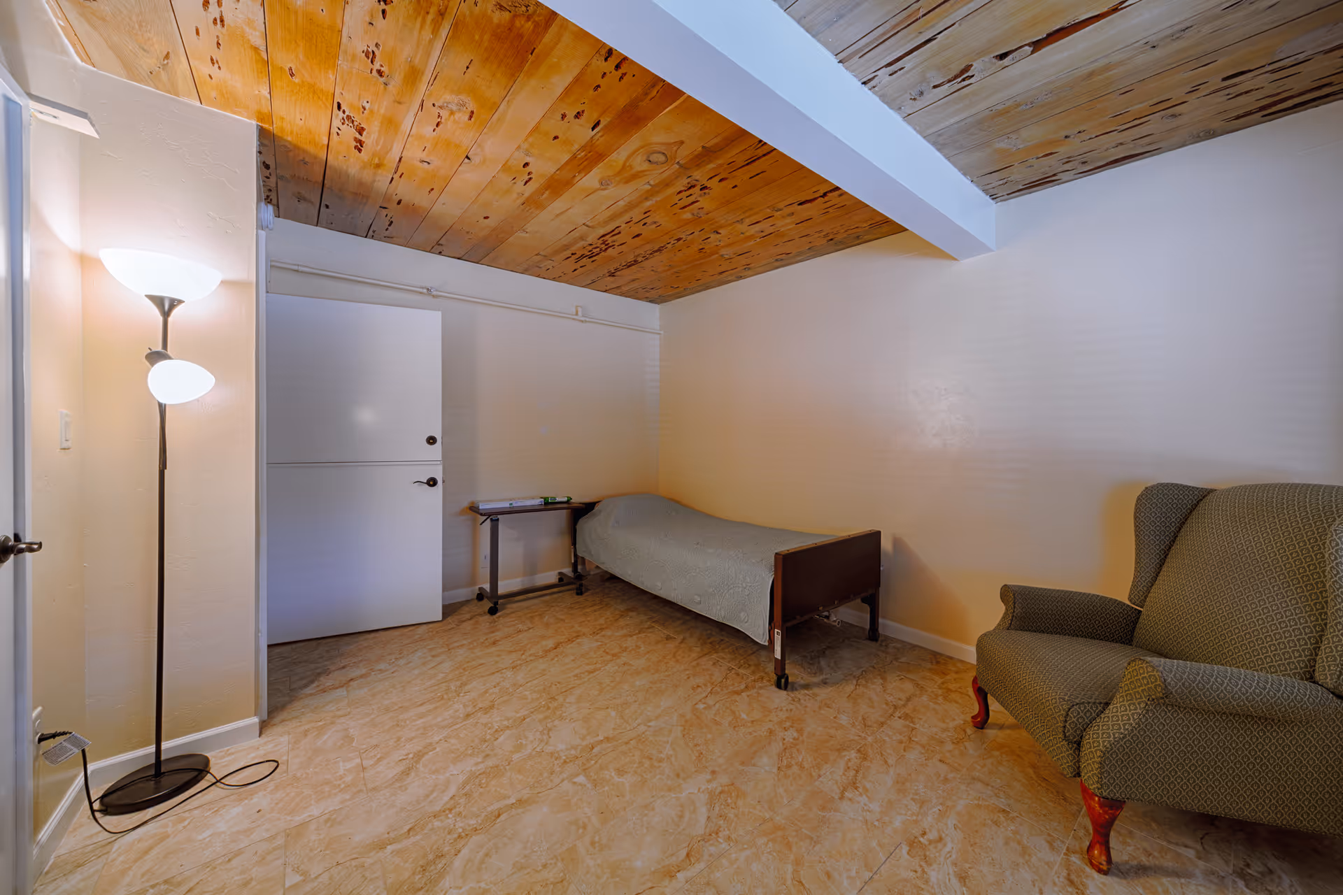 A small room with a wooden ceiling, beige walls, and tiled floor. The room contains a single bed with a gray blanket, a small table with some items on it, a green upholstered armchair with wooden legs, and a tall floor lamp with two lights. There is a white door on the left side of the room.