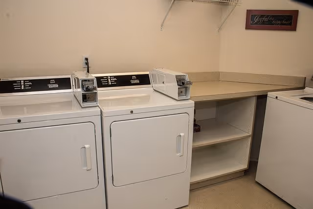 Laundry room with two white coin-operated dryers side by side, a countertop with open shelving underneath, and a washing machine partially visible on the right. A small framed sign on the wall reads 'Joyful living begins here.'