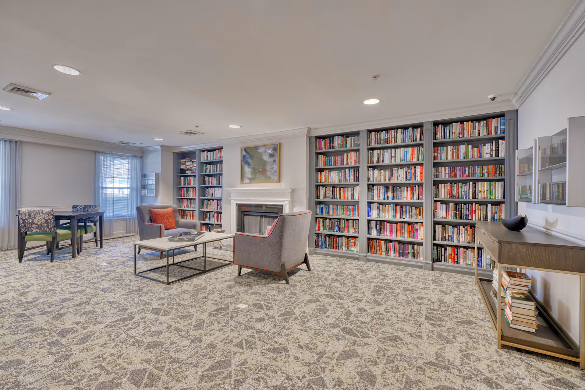 Cozy common room with wall-to-wall bookshelves, a fireplace, armchairs, a coffee table and additional seating near windows.