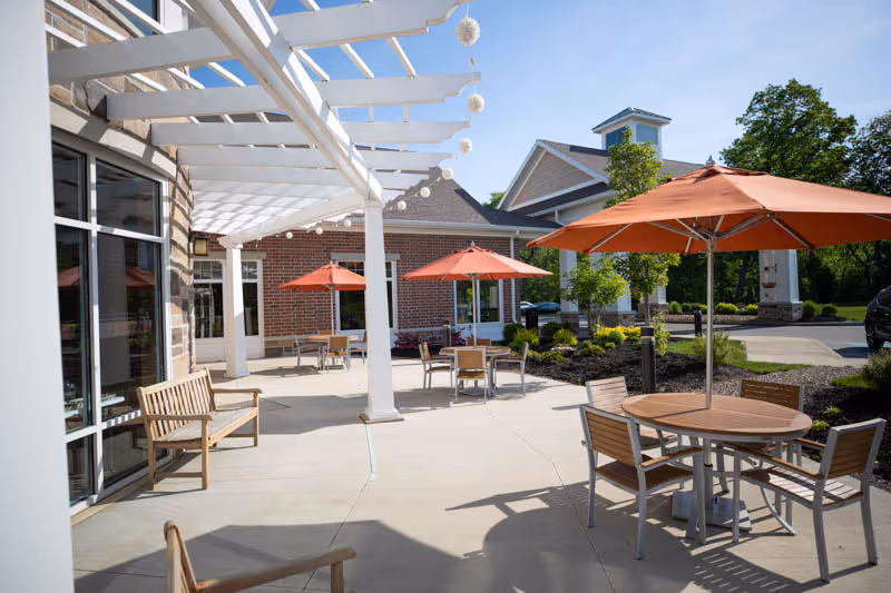 Outdoor patio with round tables, orange umbrellas, wooden benches, and a white pergola beside a brick building.