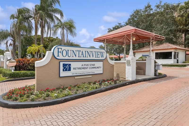 Entrance sign for Fountainview, a five star retirement community, with a gated entry booth and landscaped flower bed under a partly cloudy sky.