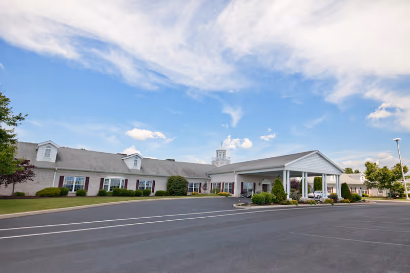 Exterior view of a single-story retirement community building with a covered entrance, surrounded by neatly trimmed bushes and a large paved parking area under a partly cloudy blue sky.