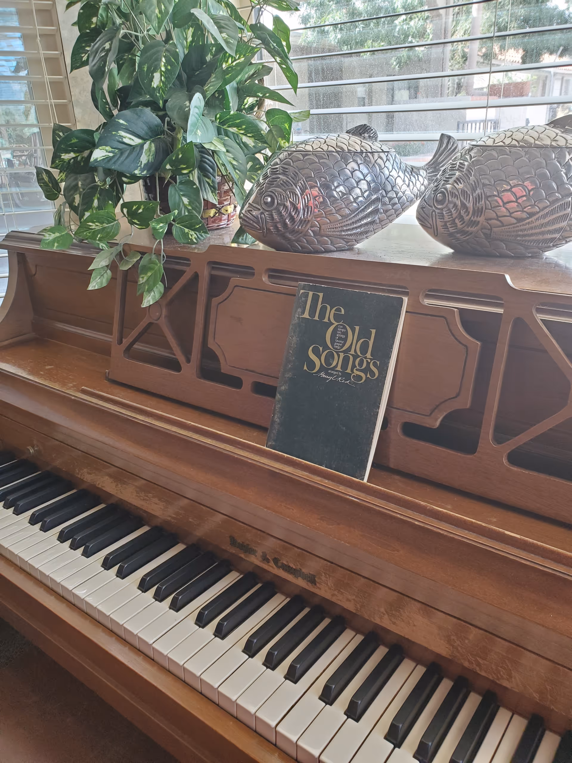 Upright wooden piano with sheet music titled 'The Old Songs', decorative fish sculptures and a potted plant on top in front of a window with blinds.