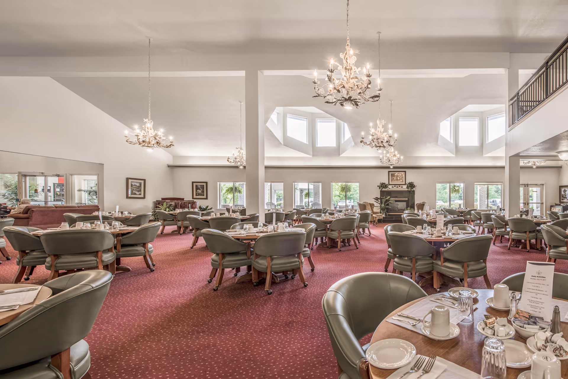 Large dining room with multiple round tables set for meals, green chairs, chandeliers, and skylights.