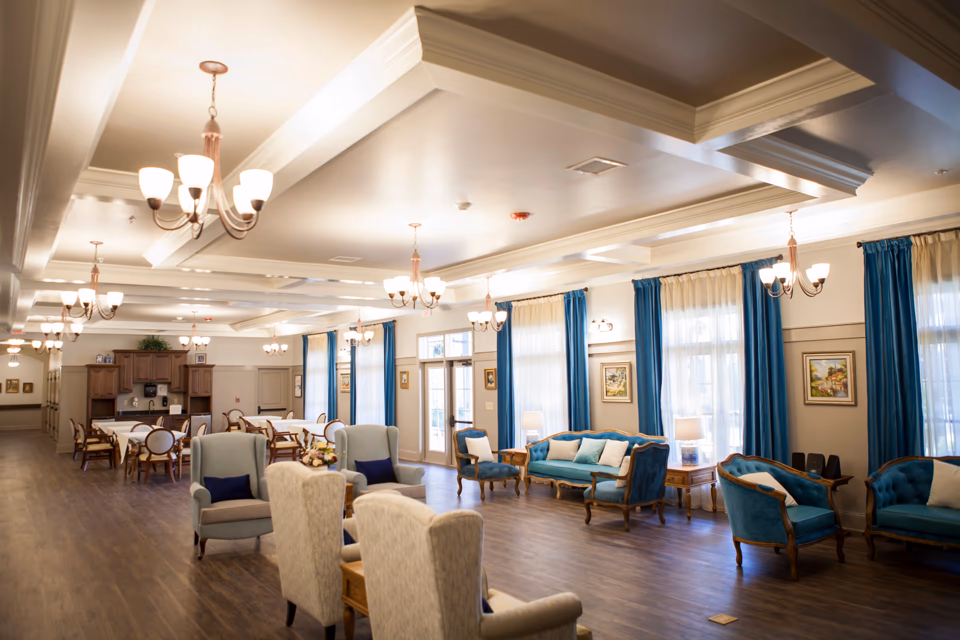 A spacious, well-lit common area in a senior living facility featuring multiple seating arrangements with upholstered chairs and sofas in blue and beige tones. The room has large windows with blue curtains, wooden flooring, and several chandeliers hanging from a coffered ceiling. In the background, there are dining tables and chairs near a wooden cabinet and a door leading outside.