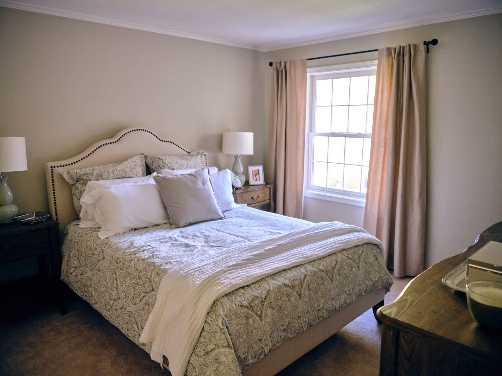 A cozy bedroom with a neatly made bed featuring patterned bedding and multiple pillows. There are two bedside tables with lamps on each side of the bed. A window with beige curtains allows natural light into the room. A wooden dresser is partially visible on the right side.