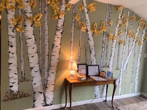 Small wooden table with a lamp and decorative items placed against a green wall painted with birch trees and yellow leaves.