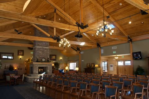Large wood‑beamed community room with rows of chairs facing a stone fireplace under a high vaulted ceiling.