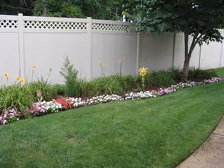A well-maintained garden area with a green lawn, a flower bed containing various colorful flowers, and a white privacy fence in the background with some trees providing shade.