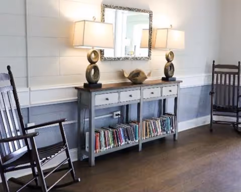 A sitting area with a console table filled with books, two lamps and a mirror on the wall, flanked by wooden rocking chairs.