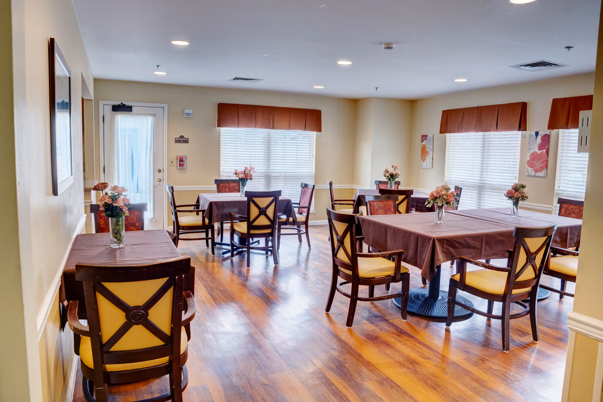 A bright dining room with several tables covered with brown tablecloths, each adorned with a vase of flowers. The room has wooden floors, yellow walls, large windows with blinds and orange valances, and wooden chairs with yellow cushions.