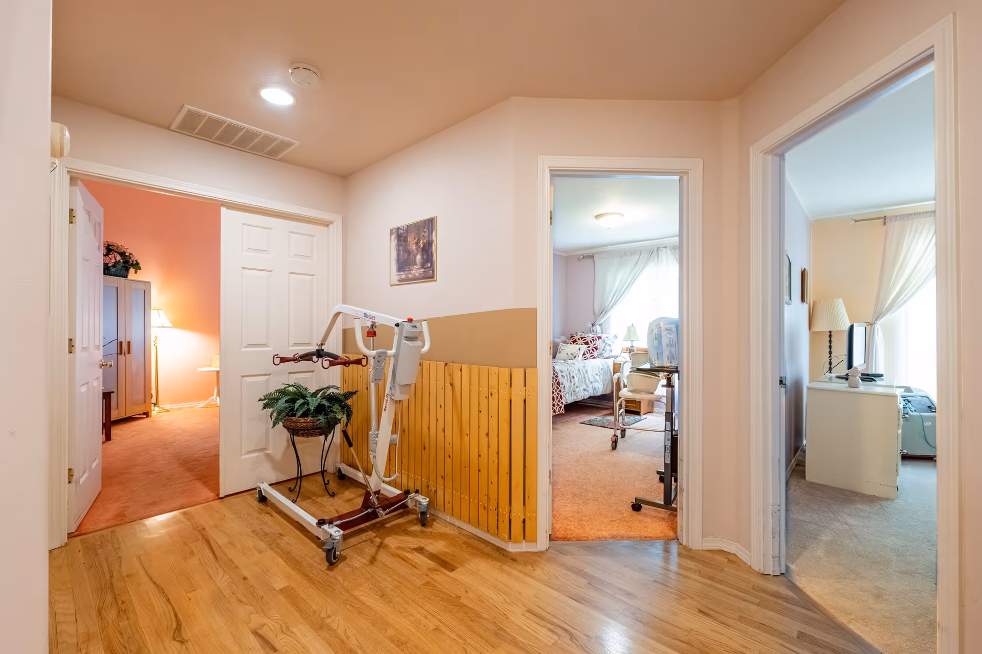 Interior hallway of a senior care facility with wooden flooring and light-colored walls. There is a patient lift device and a potted plant against the wall. Three doorways lead to different rooms, each with carpeted floors and furnished with beds, lamps, and other furniture. Soft natural light filters through curtains in the rooms.