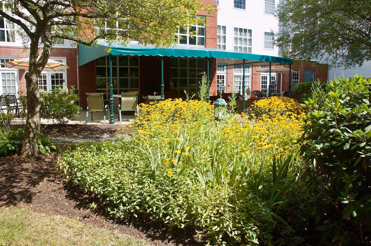 Outdoor garden area with yellow flowers and green shrubs in front of a brick and white building. There is a covered patio with green awning and outdoor chairs and tables. A person is sitting on the patio.