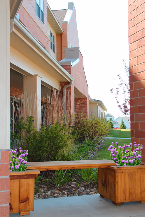 Outdoor view of a senior living facility showing a wooden bench with planters containing purple flowers on each side. The bench is placed on a concrete path next to a landscaped garden with bushes and tall grasses. The building exterior features brick and siding with windows and a sloped roof.