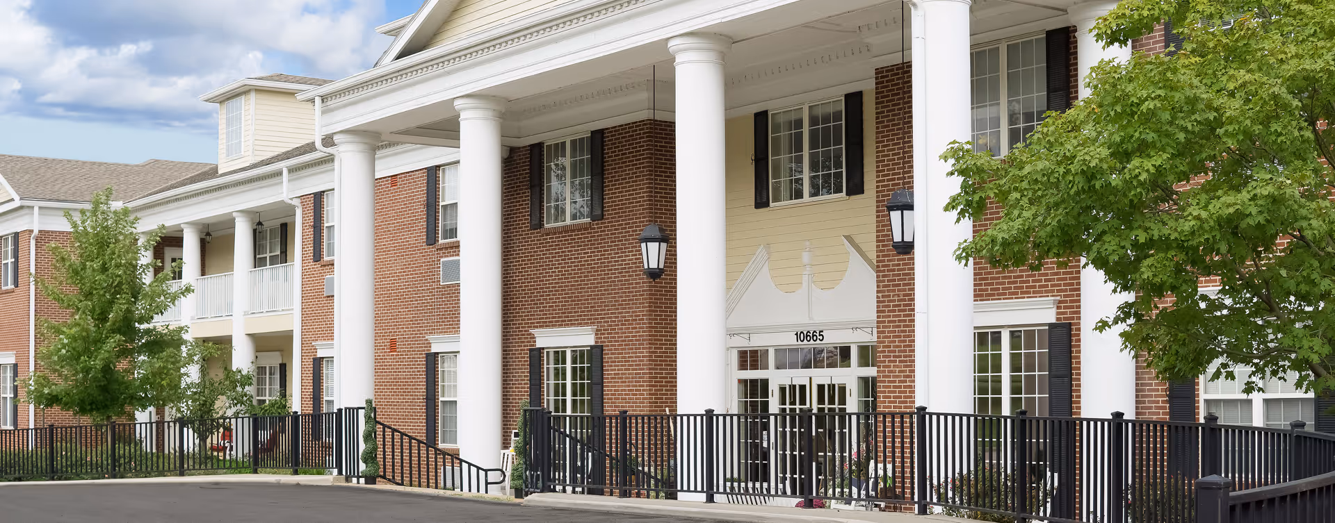 Exterior view of a senior living facility with red brick walls, large white columns, black railings, and green trees under a partly cloudy sky.