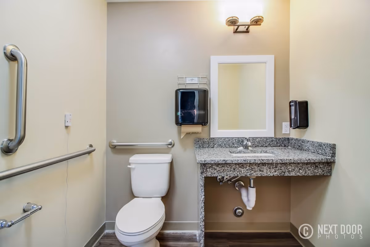 A clean and accessible bathroom with a white toilet, granite countertop with an under-mount sink, a rectangular mirror above the sink, and safety grab bars on the walls. There are a paper towel dispenser and a soap dispenser mounted on the walls. The floor has wood-like vinyl flooring.