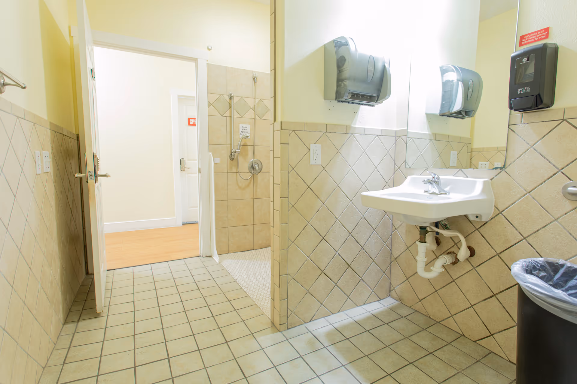 A clean, tiled bathroom with a wall-mounted sink, two paper towel dispensers, a soap dispenser, and a trash can. There is an open door leading to a hallway with wooden flooring. The shower area is visible with a handheld showerhead and a small sign on the wall.