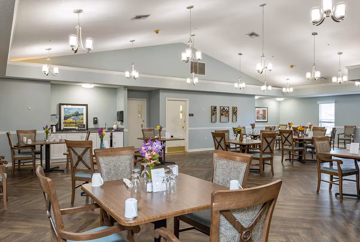 A spacious dining room with multiple wooden tables and chairs arranged neatly. Each table has a small flower vase with colorful flowers, glassware, and white napkins. The room has light blue walls, wooden flooring, and several modern hanging light fixtures. There are framed pictures and decorative wall art on the walls, and a small counter area with a coffee machine and water dispenser in the background.