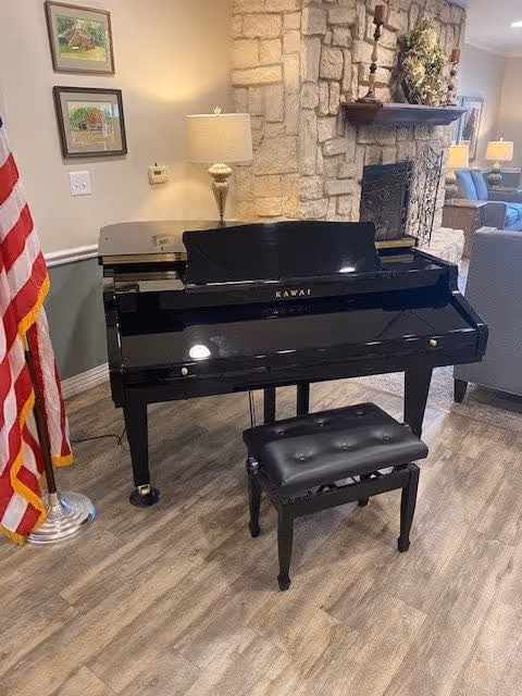 A black Kawai grand piano with a matching black cushioned bench is placed on a wood floor in a cozy room. To the left of the piano is an American flag on a stand. Behind the piano is a stone fireplace with a wooden mantel decorated with a floral arrangement and candle holders. A table lamp and framed pictures hang on the wall to the left. In the background, there is a gray armchair and another lamp.