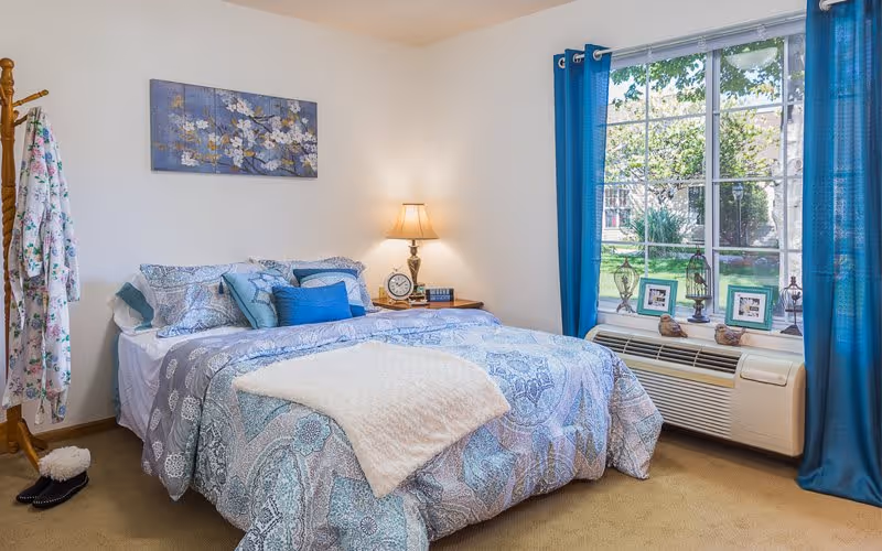 A cozy bedroom with a neatly made bed featuring blue and white patterned bedding and multiple pillows. A wooden nightstand with a lamp, clock, and books sits beside the bed. A large window with blue curtains lets in natural light and overlooks a green outdoor area. A wooden coat rack with a floral robe and slippers is positioned near the bed.