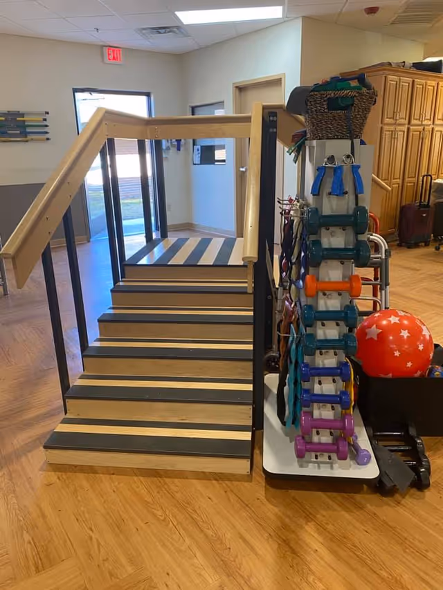 Indoor physical therapy area with a wooden staircase equipped with handrails on both sides and a rack holding various colorful dumbbells, resistance bands, and exercise balls. The room has wooden flooring and light-colored walls with a door and exit sign visible in the background.