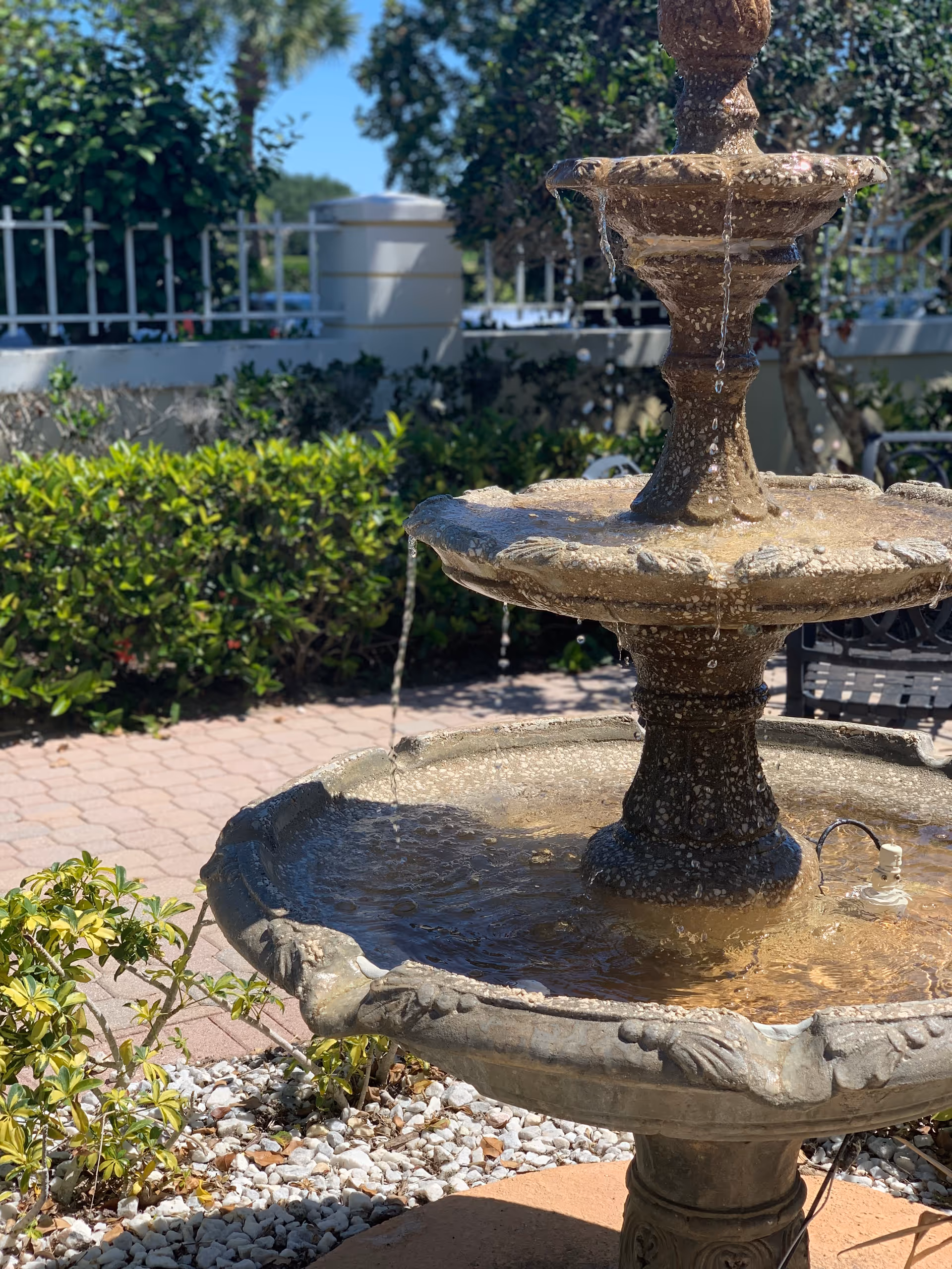 A close-up view of a multi-tiered stone water fountain with water flowing from the top tier to the lower basins, surrounded by green bushes, a white fence, and a paved patio area under a clear blue sky.