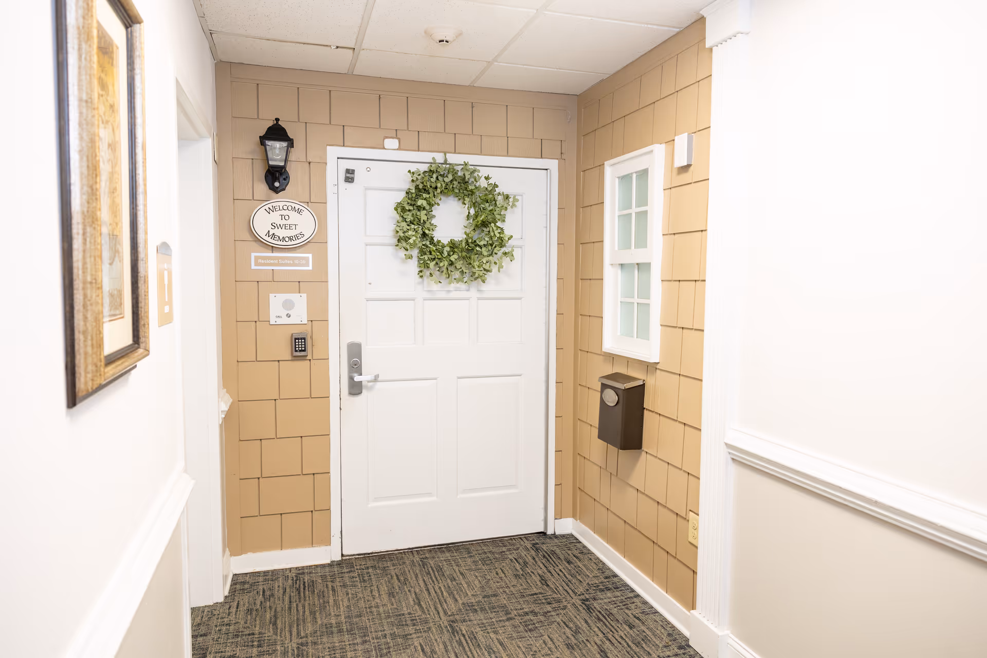 Interior hallway corner with a white door decorated with a green wreath. The walls are beige with shingle-style paneling and white trim. A small black wall lantern is mounted above a sign that reads 'Welcome to Sweet Memories' and another sign indicating 'Resident Suites 10-30'. There is a small window with white trim on the right wall and a black mailbox or drop box mounted below it. The floor is carpeted with a patterned design.