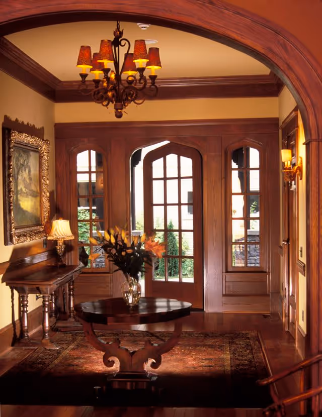 A warmly lit interior room with wooden trim and a large arched doorway. The room features a round wooden table with a vase of flowers in the center, a side table with a lamp, a framed painting on the wall, and a chandelier with multiple lampshades hanging from the ceiling. Large windows and a glass door provide a view of greenery outside.