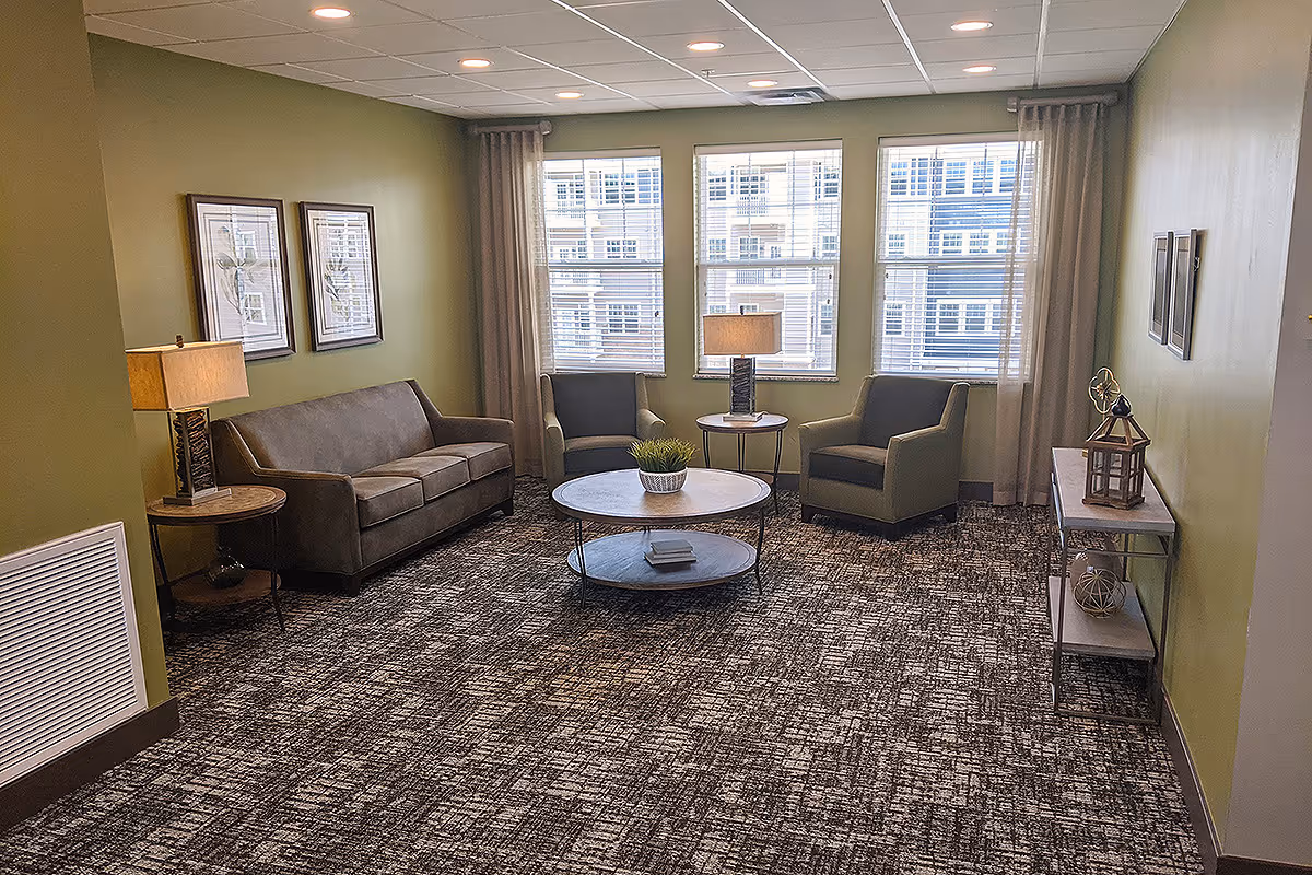 A cozy living room area with green walls and patterned carpet. The room features a brown sofa, two green armchairs, a round coffee table with a small plant, two side tables with lamps, framed botanical artwork on the walls, and a console table with decorative items. Three large windows with blinds and curtains allow natural light to enter the space.