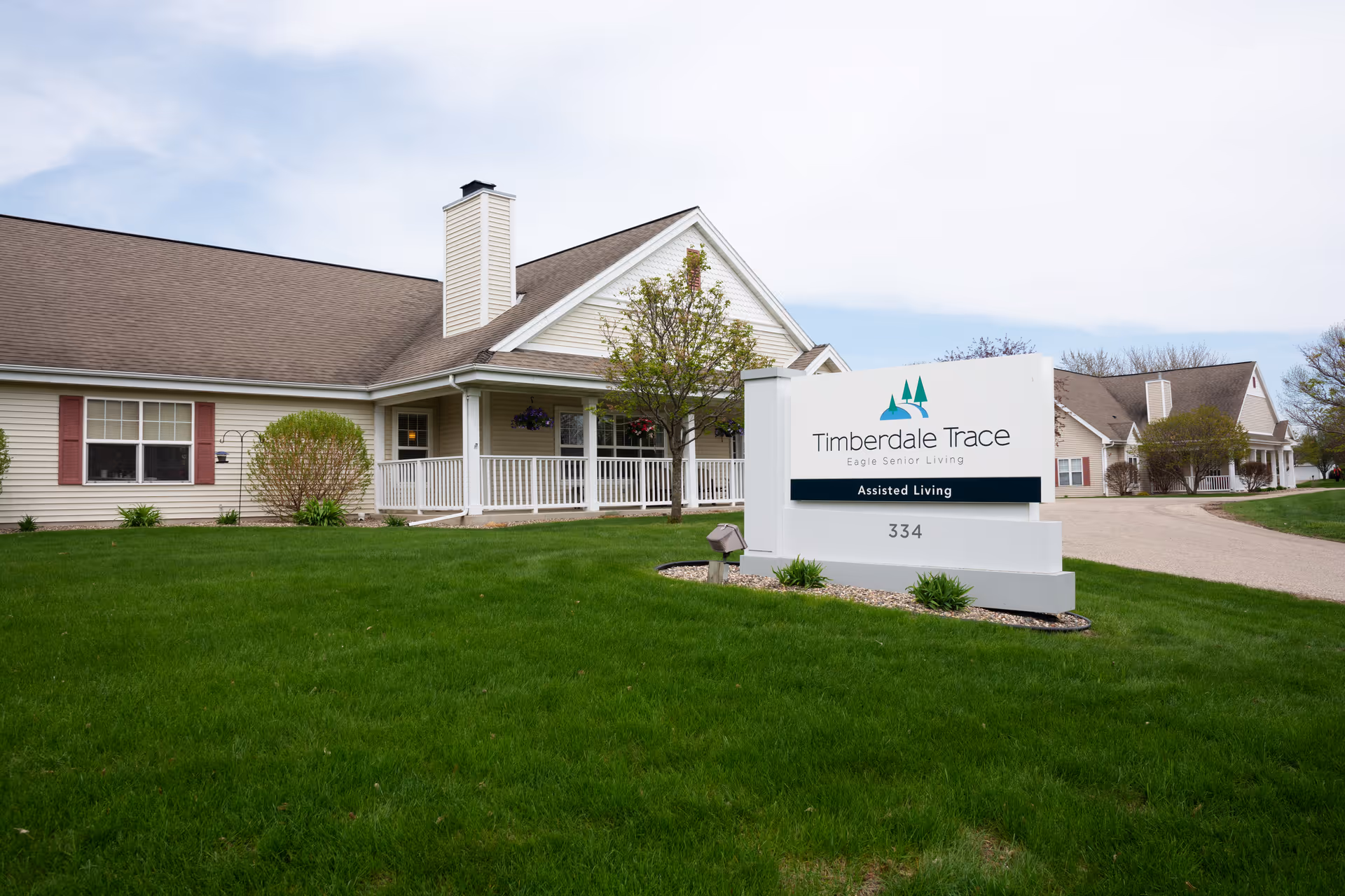 Exterior view of Timberdale Trace Assisted Living facility showing a single-story building with beige siding, white trim, and a brown roof. There is a white sign in front with the facility name and address, surrounded by green grass and small shrubs under a partly cloudy sky.
