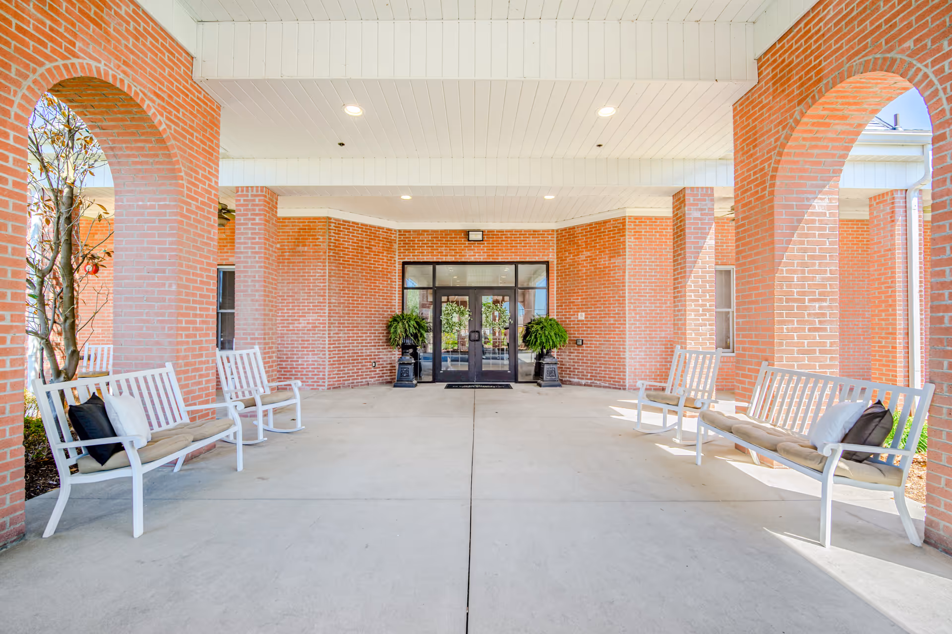 Covered entrance area of a brick building with white benches on both sides, each bench having cushions and pillows. The entrance features glass double doors with two large potted plants on either side. The ceiling is white with recessed lighting, and the area is supported by brick columns with arches.