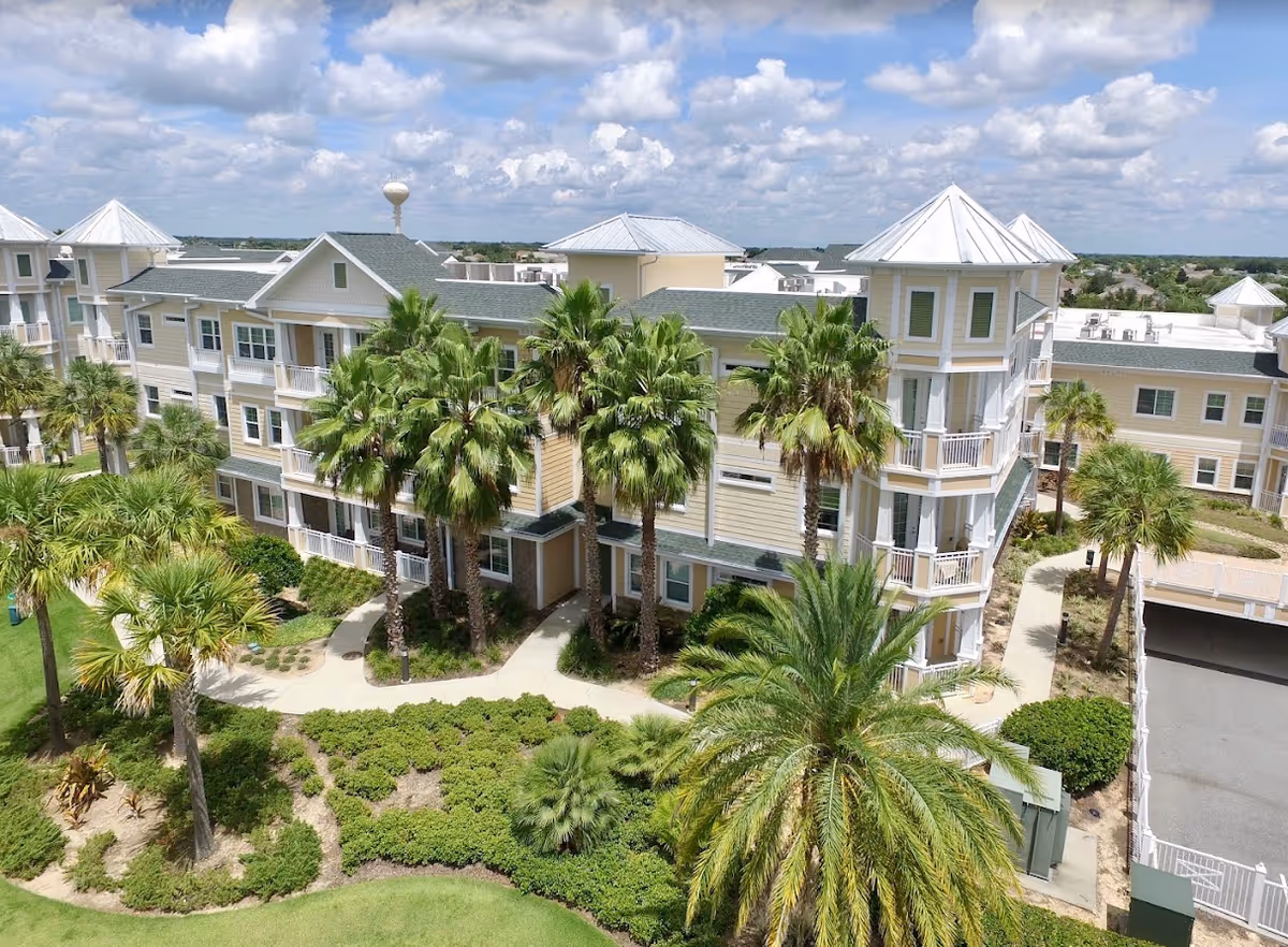 Aerial view of Sumter Place In The Villages senior living facility showing a multi-story building with beige siding and green roofs surrounded by palm trees and landscaped greenery under a partly cloudy sky.