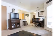 Bright open living and dining area with a TV on a wooden console, a round dining table with chairs, and bar stools at a kitchen counter.