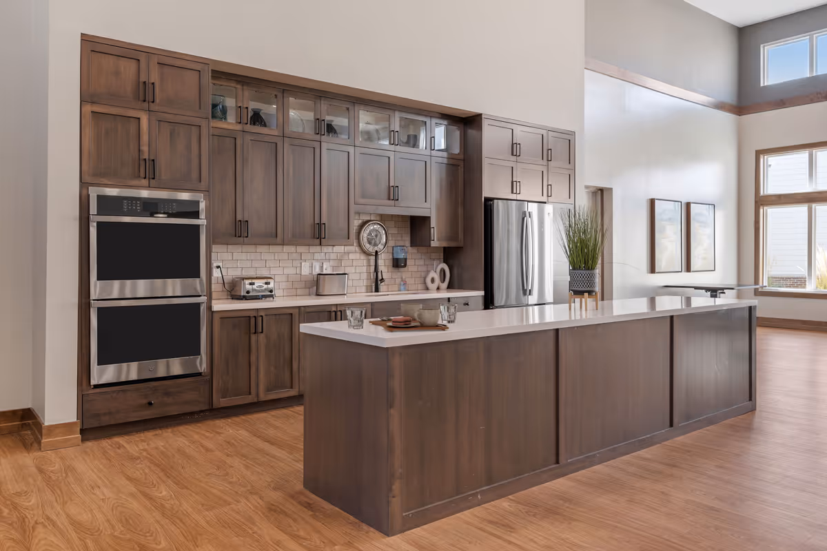 A modern kitchen with wooden cabinets and a large island countertop. The kitchen features stainless steel double ovens, a refrigerator, a toaster, and a coffee maker. There is a plant on the island and two framed pictures on the adjacent wall. The room has large windows allowing natural light to fill the space.