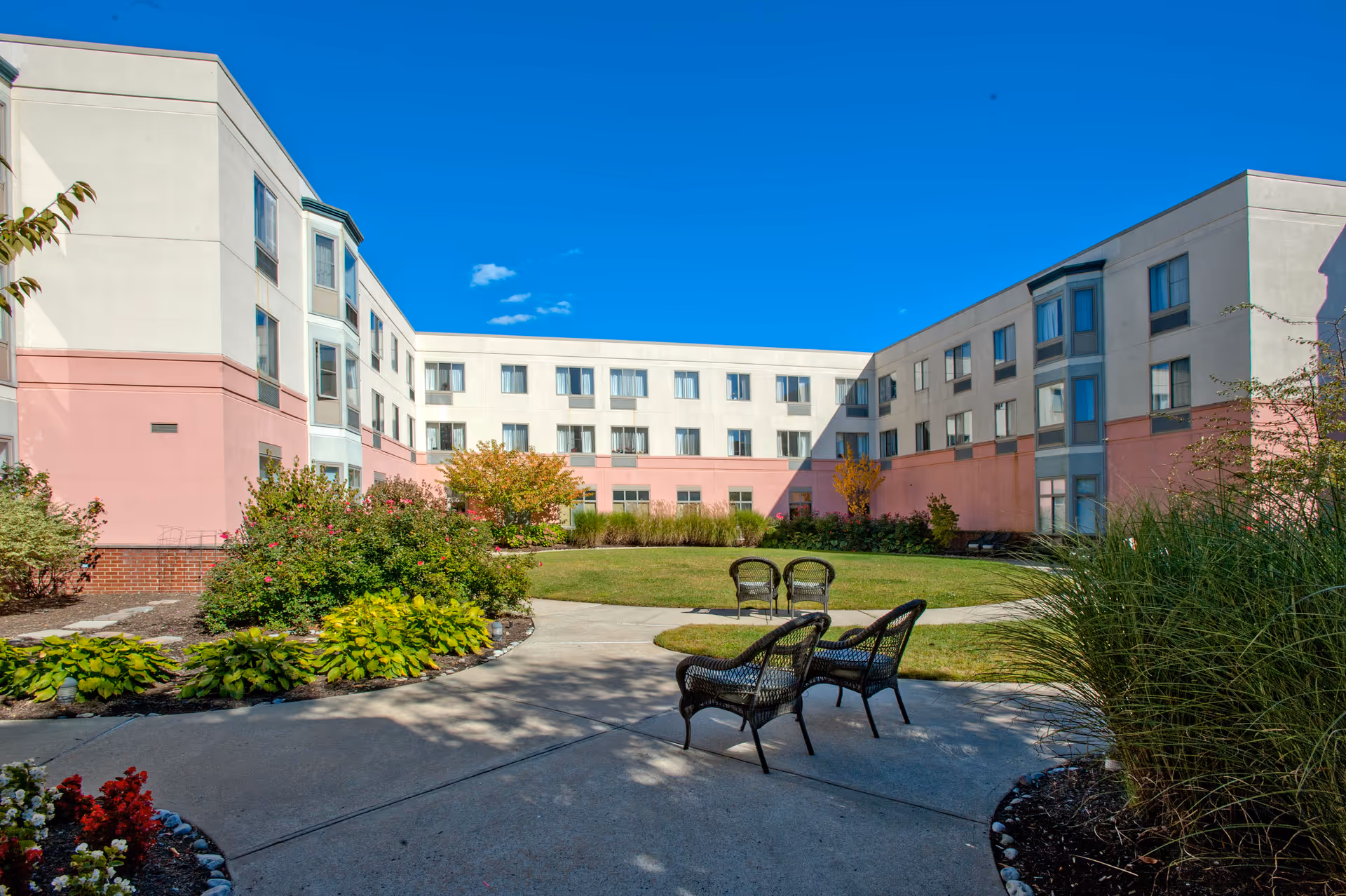 Courtyard with patio chairs, lawn and gardens surrounded by a three-story pink-and-white senior living building under a clear blue sky.