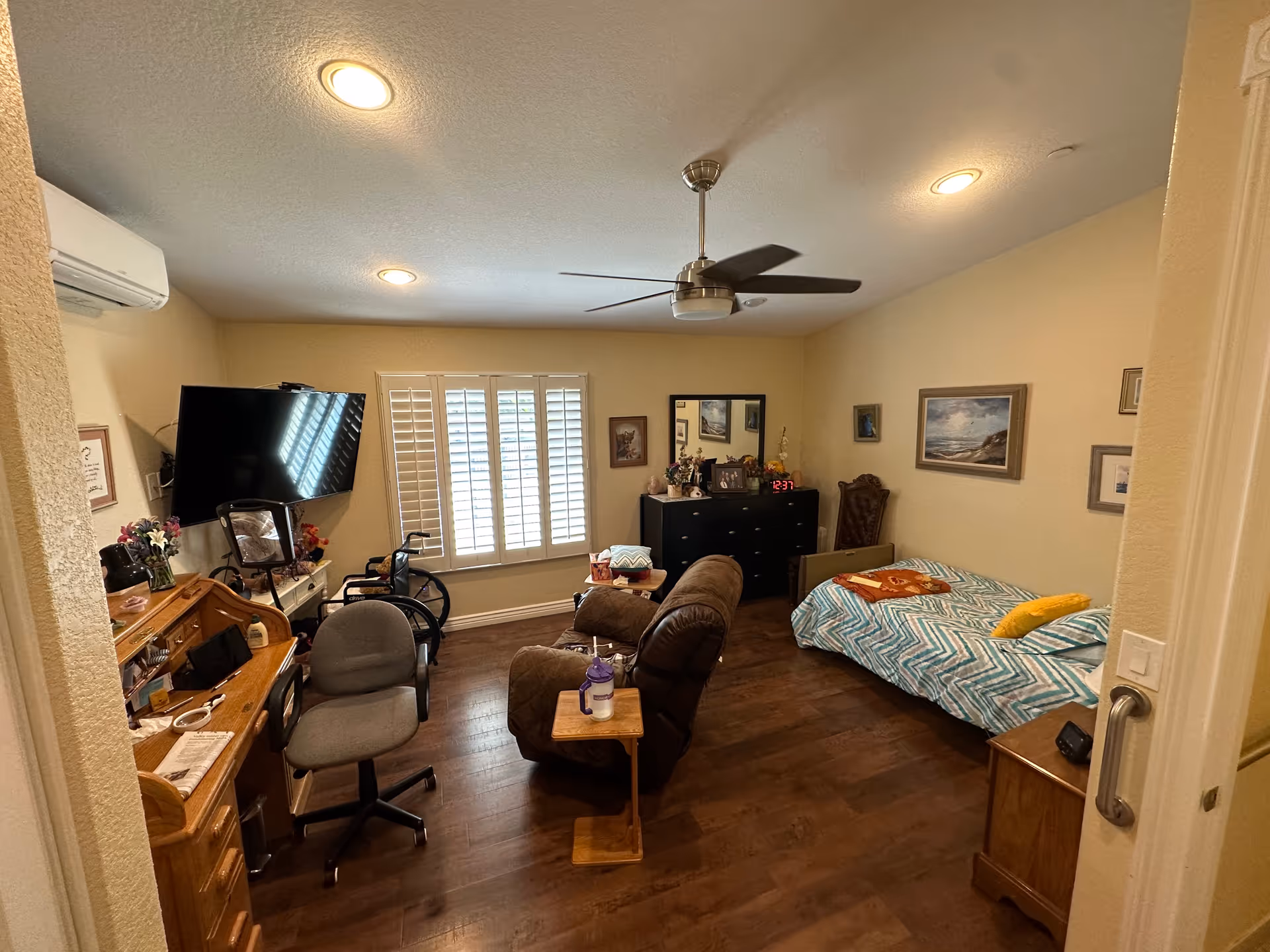 A cozy assisted living room with a single bed covered in a blue and white chevron patterned blanket, a brown recliner chair with a small wooden side table, a gray office chair, a wheelchair, a wall-mounted flat screen TV, a wooden desk with various items, a black dresser with a mirror and decorative items, and several framed pictures on the walls. The room has wooden flooring, a ceiling fan, and a window with white shutters.