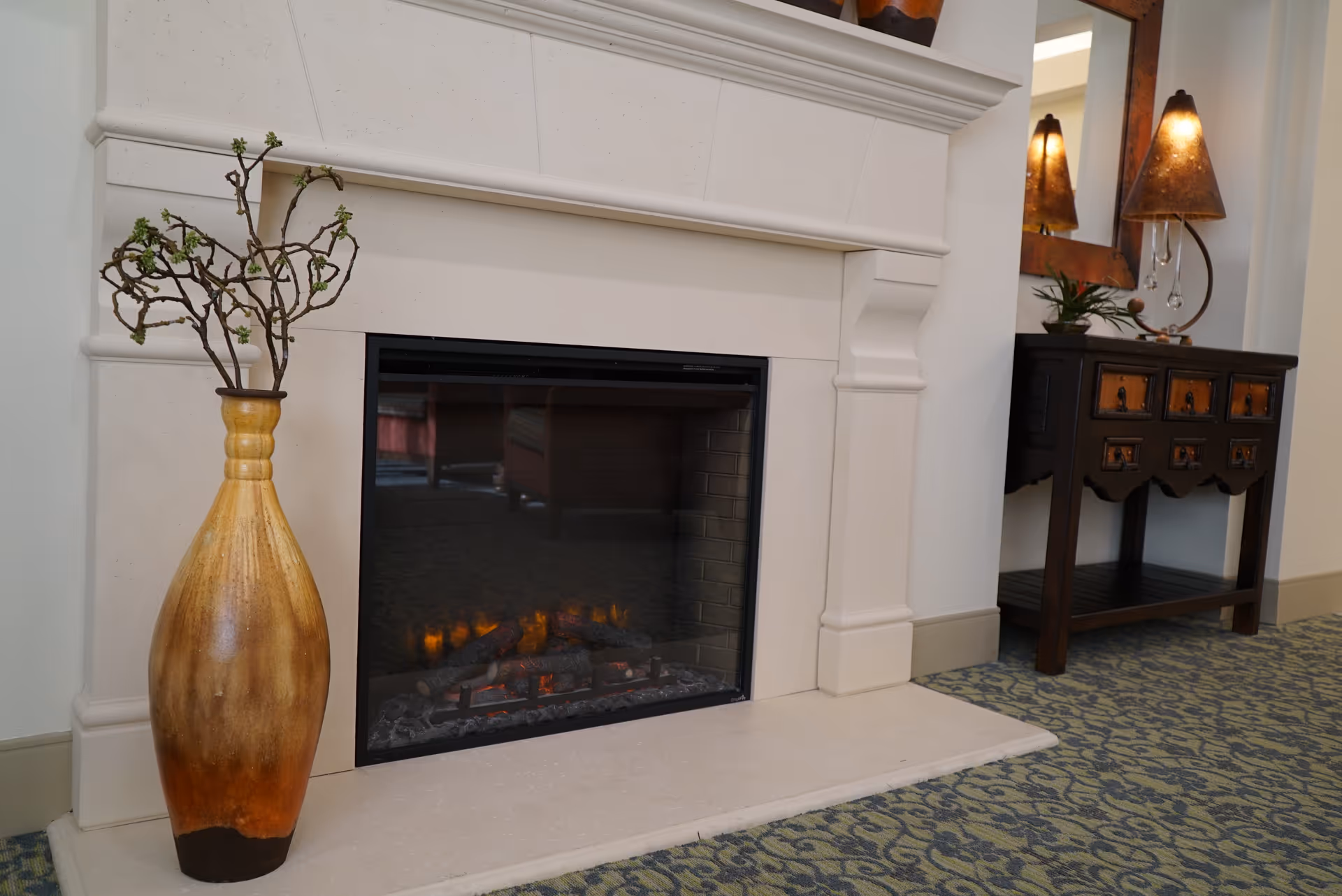 A cozy indoor scene featuring a modern electric fireplace with a beige stone mantel. To the left of the fireplace is a tall decorative vase with branches. To the right, there is a dark wooden console table with drawers, a mirror above it, and two lit lamps with brown lampshades. The floor is covered with a patterned carpet in shades of green and blue.