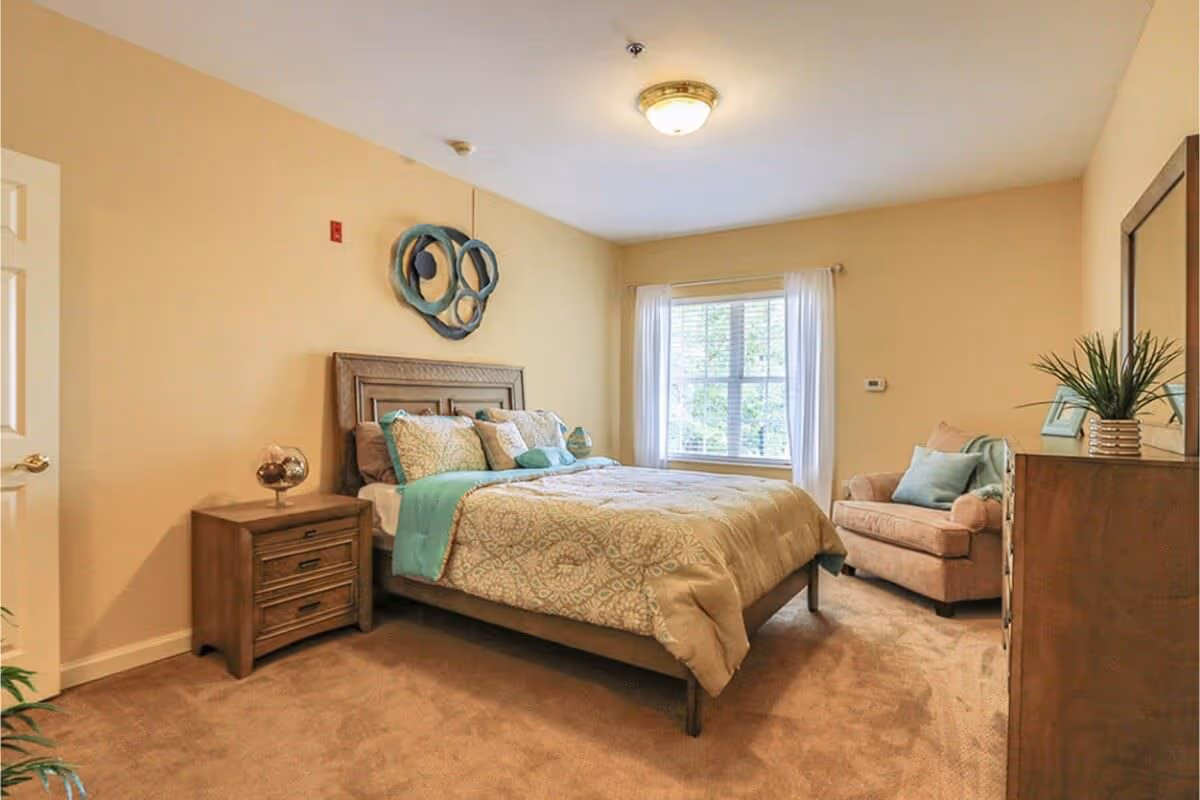 A cozy bedroom with beige walls and carpet, featuring a wooden bed with patterned bedding and teal accents, a matching wooden nightstand with a decorative lamp, a large window with white curtains, a comfortable armchair with pillows, and a wooden dresser with a mirror and a potted plant.
