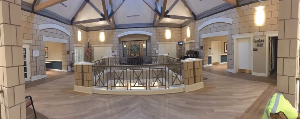 Interior view of a senior living facility with a spacious common area featuring high vaulted ceilings with exposed beams, beige brick-patterned walls, wooden flooring, and a central railing around an open lower level. There are multiple doorways leading to other rooms, including one labeled 'Grandview Theater'. An American flag is displayed near one doorway, and chairs are arranged along the walls.