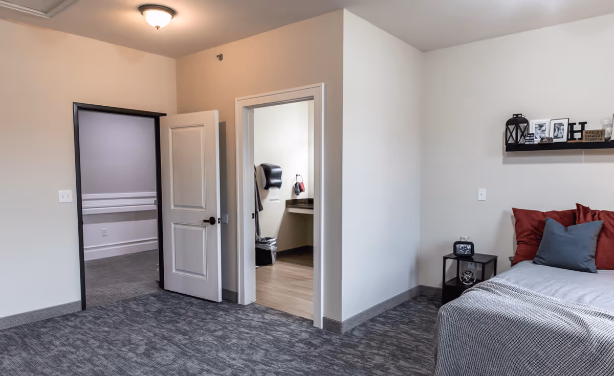 Interior view of a senior living facility room with a bed on the right side adorned with red and gray pillows and a gray blanket. A small black nightstand next to the bed holds a clock and decorative items. On the wall above the bed is a shelf with framed photos and decorative objects. Two open doorways are visible, one leading to a hallway and the other to a bathroom with a paper towel dispenser and a trash can. The room has gray carpet and beige walls with a ceiling light fixture.
