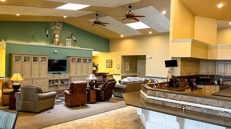A spacious and well-lit common area in an assisted living facility featuring a seating arrangement with armchairs and sofas around a large entertainment center with a TV. The room has ceiling fans, decorative items on top of the cabinets, and a kitchen counter with appliances visible on the right side.