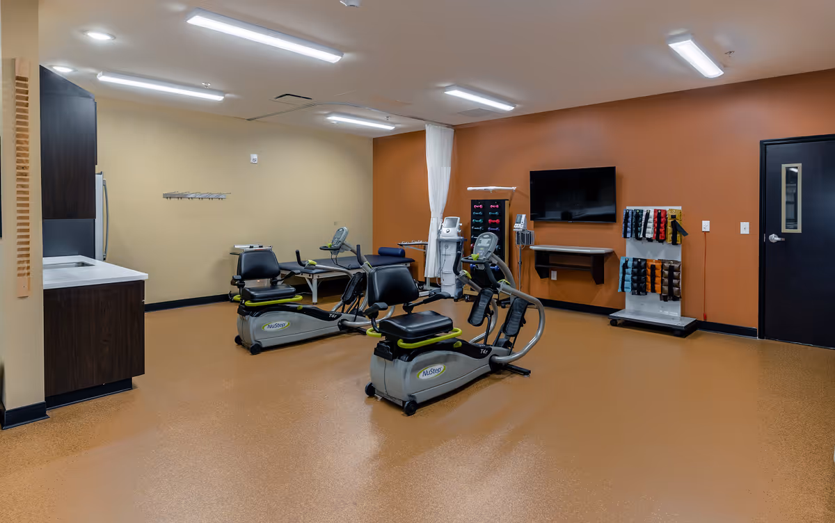 A fitness room with two NuStep exercise machines, a small sink and cabinet area on the left, a wall-mounted TV, and a rack holding various colored resistance bands. The walls are painted beige and orange, and the floor has a brown speckled surface.